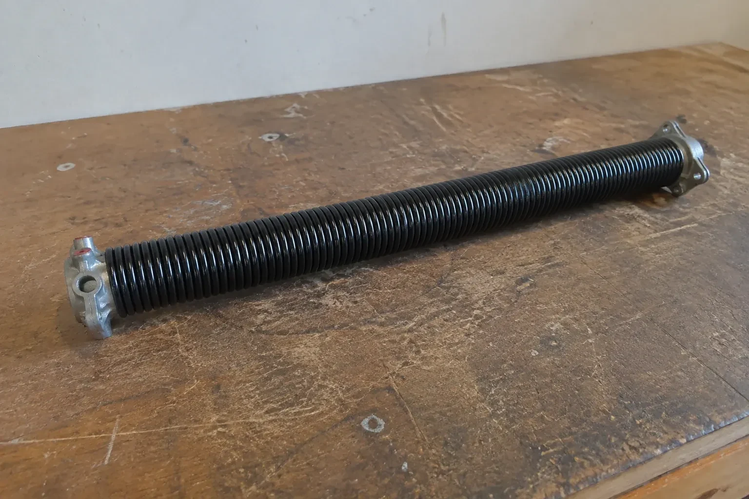 Detached black coil spring with metal end fittings on a workbench. — Torsion Spring Replacement, Oakville