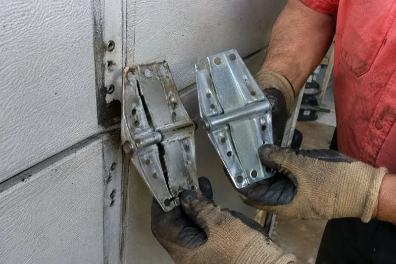 Gloved hands holding two metal garage-door hinges side by side, one discolored with a visible crack and the other bright silver. — Garage Door Hinge Replacement, Oakville